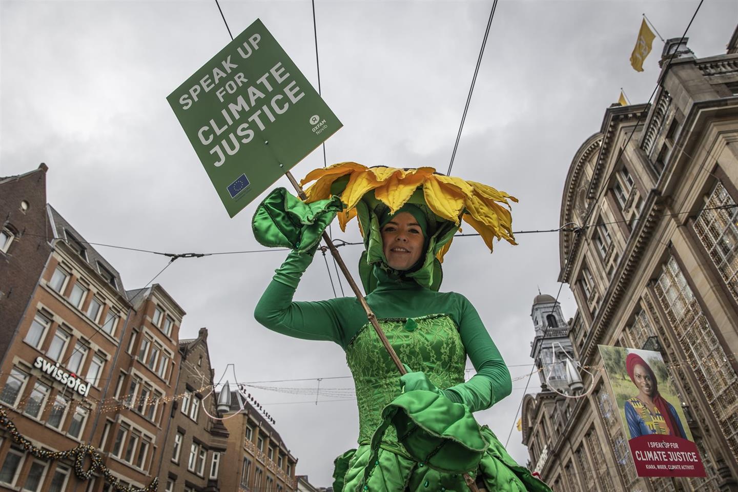 Een vrouw in een zonnebloemkostuum met een protestbord 'Speak up for Climate justice'
